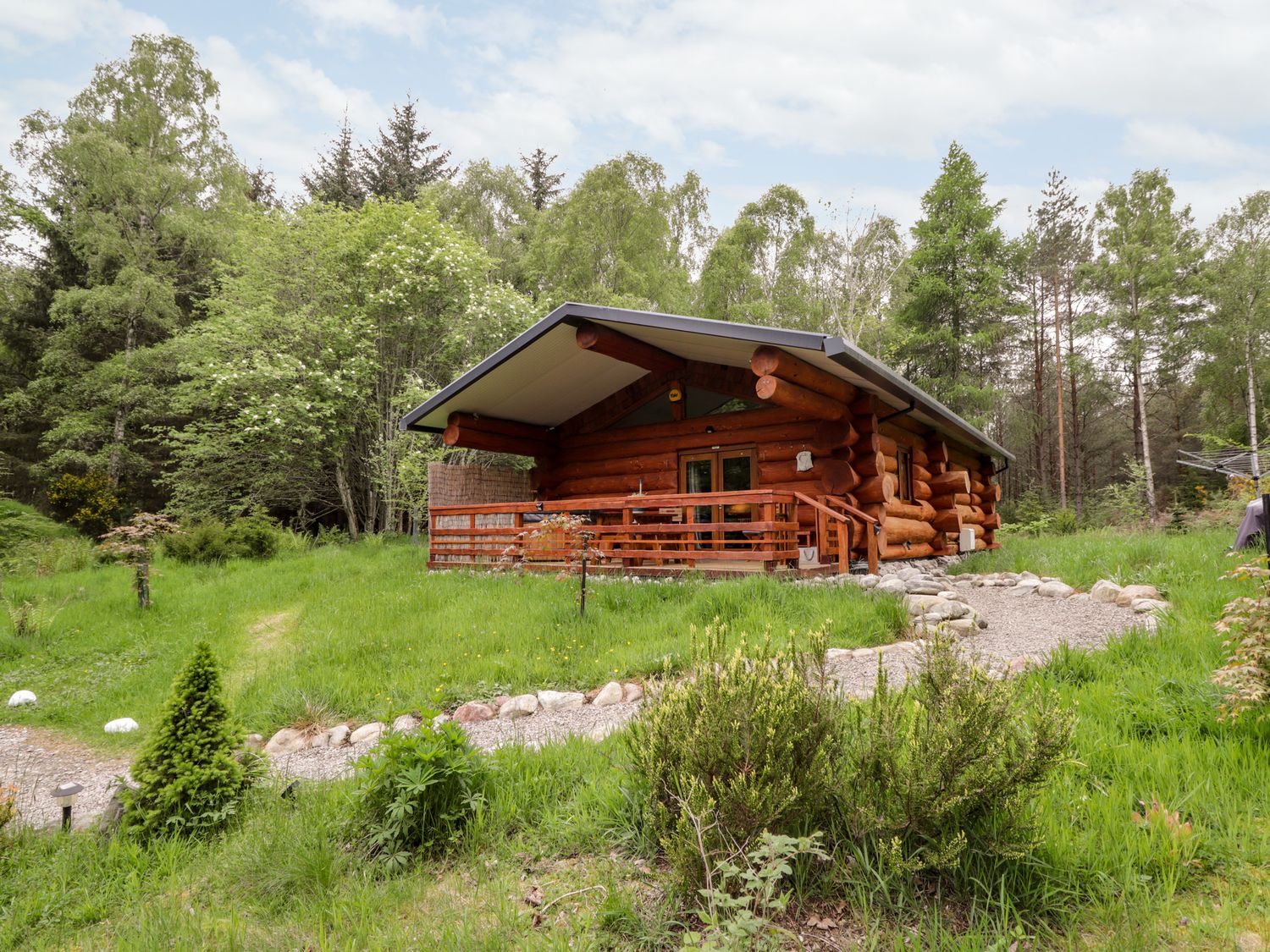 A wooden log cabin with a covered porch and a gravel pathway surrounded by grass and trees at Heron Lodge in North Kessock