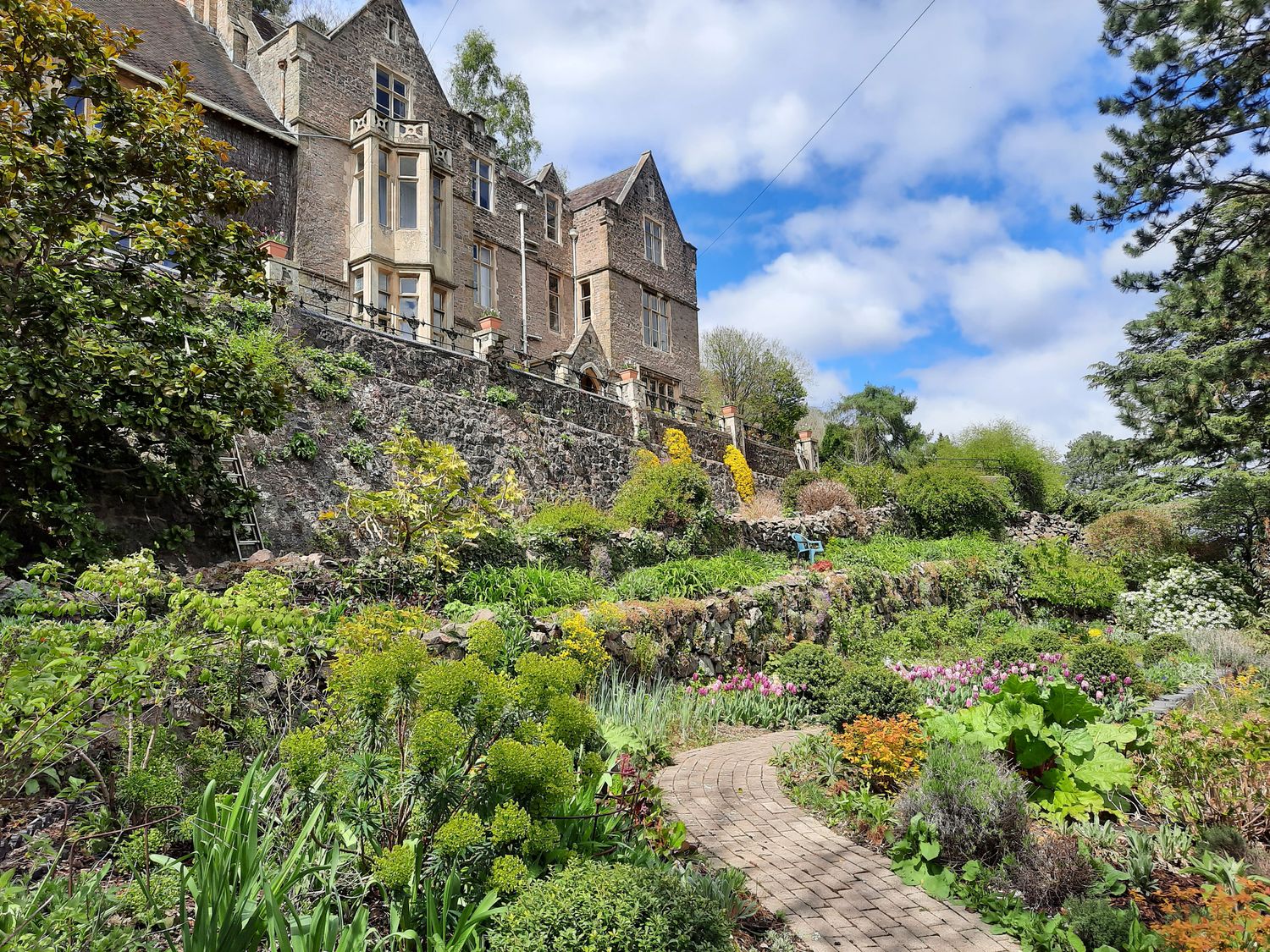 A garden with a curved brick pathway and stone terraces in front of a large stone house at Haslington House in Malvern