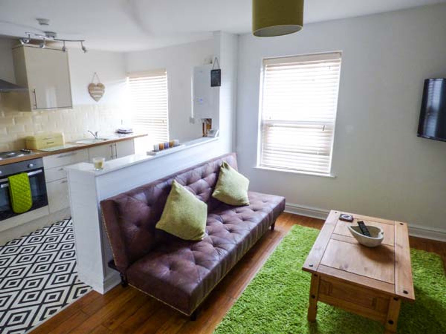 A living room with a brown sofa with green pillows a wooden coffee table a green rug and a kitchen area at Flat 3 in Bridlington