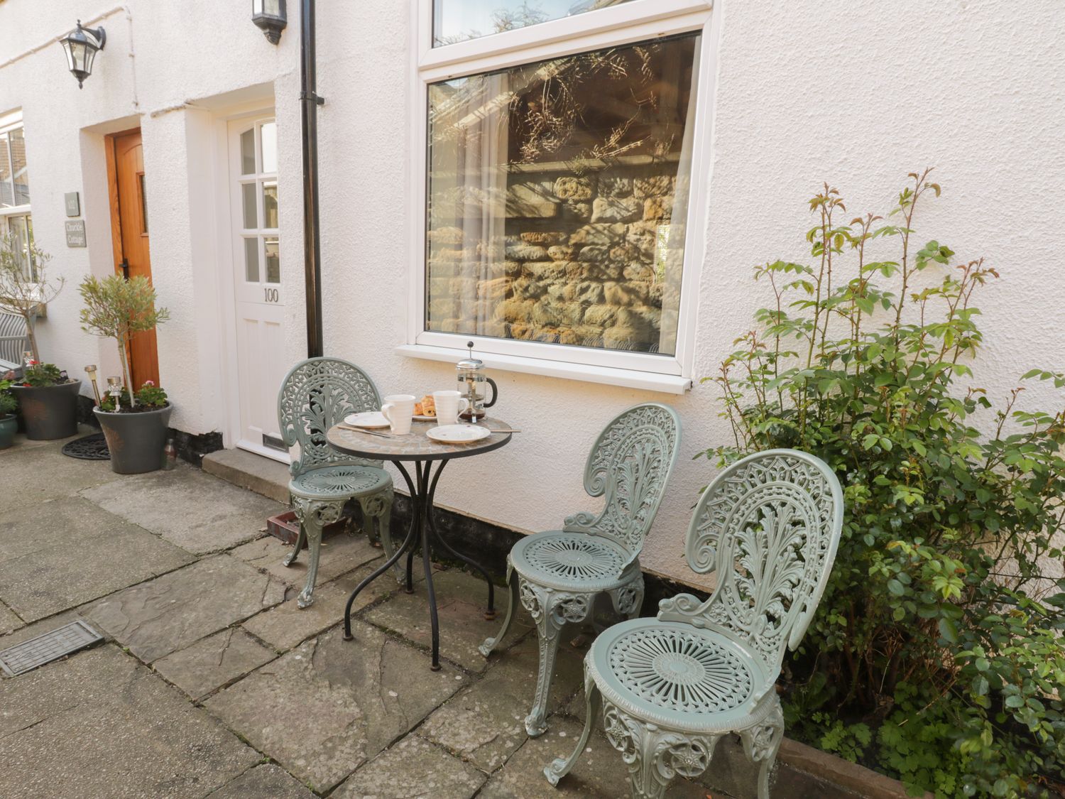 A small outdoor seating area with three metal chairs and a round table with cups and a French press outside a white building at Cosy Cottage in Swainby