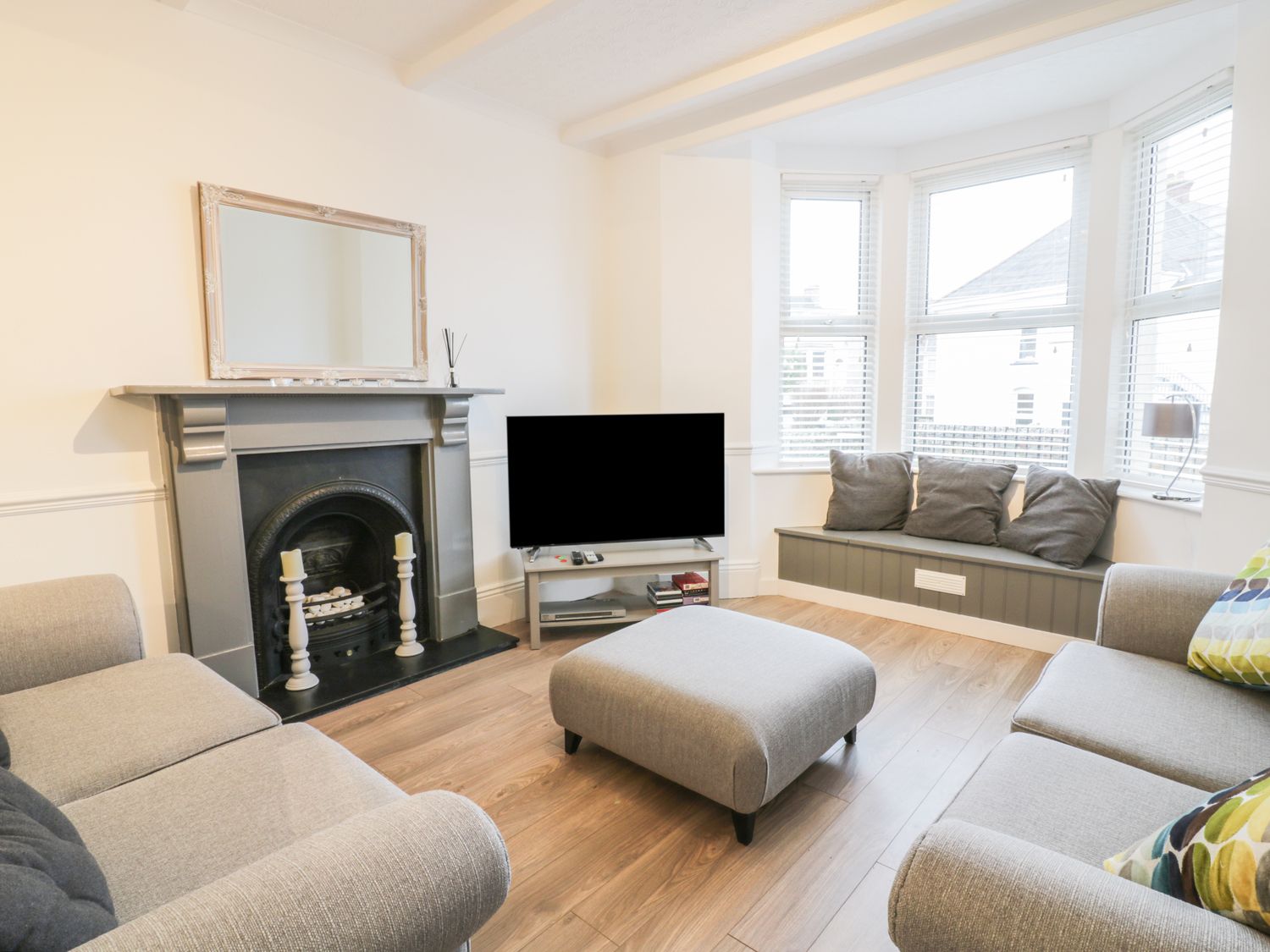 A living room with a grey fireplace a television two sofas an ottoman and a window seat with cushions at The Salt House in Newquay