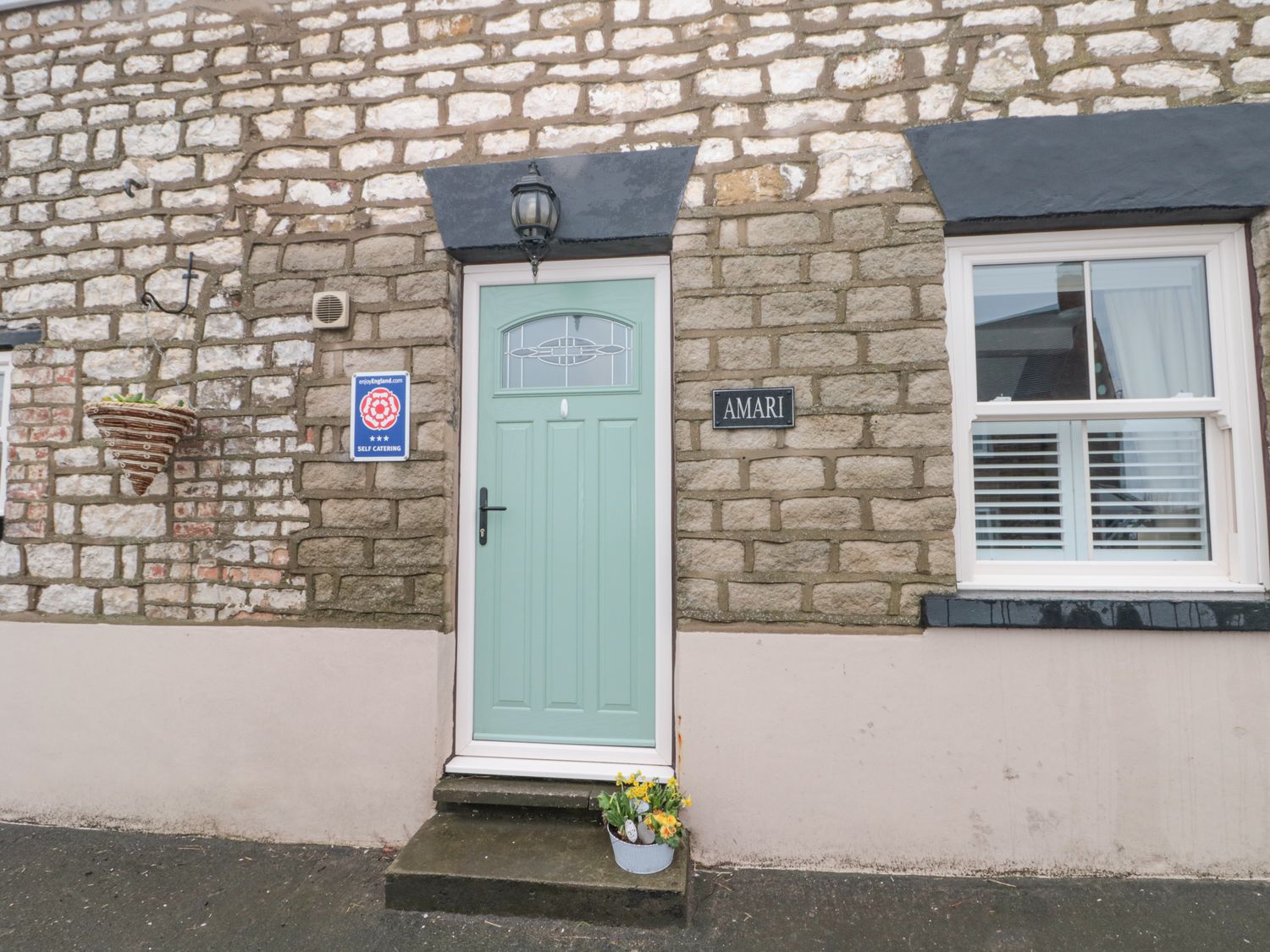 A green door with a window and a black handle on a stone house wall with a flower pot and a black sign reading Amari at Amari in Staxton
