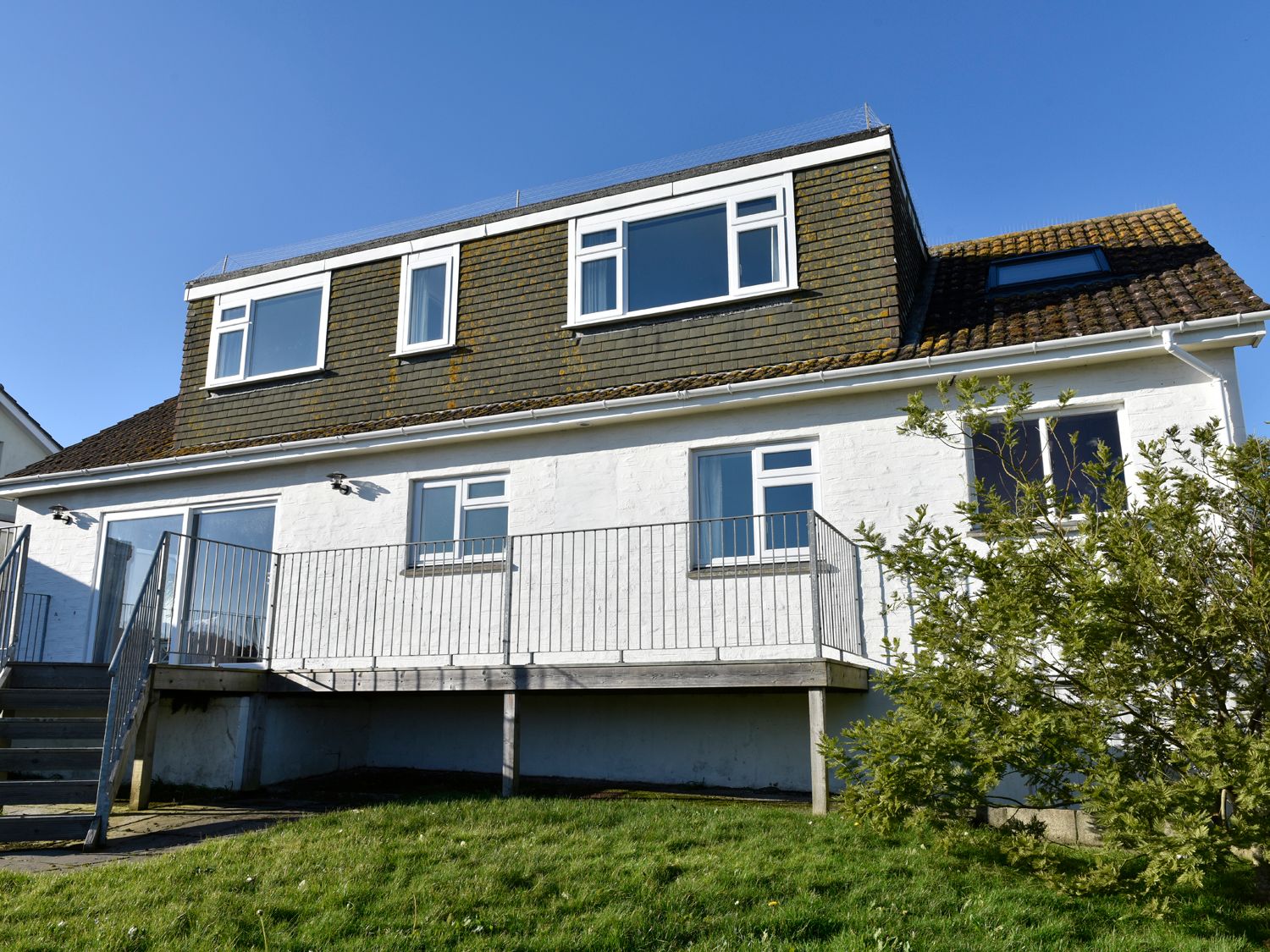 A two-story house with white walls and shingled upper story with a raised patio and metal railing at Treverrion in Gorran Haven