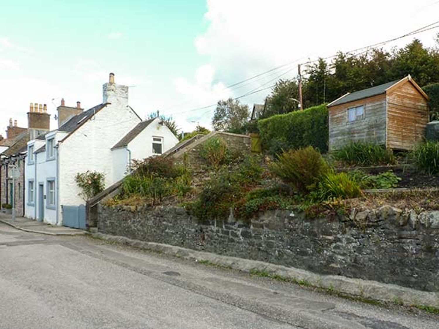 A row of white houses along a street with a stone wall and garden with shrubs and a wooden shed at Nathaniel's Cottage in Kirkcudbright