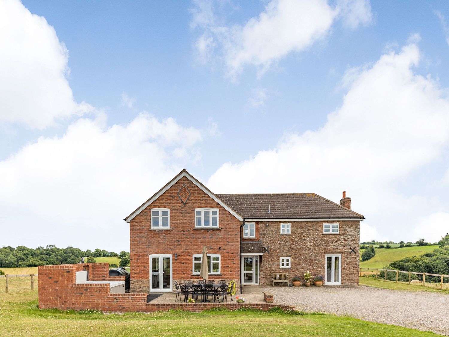 An outdoor view of a two-story house with brick and stone walls a patio with table and chairs and surrounding lawn at Wood Cottage in Tenbury Wells