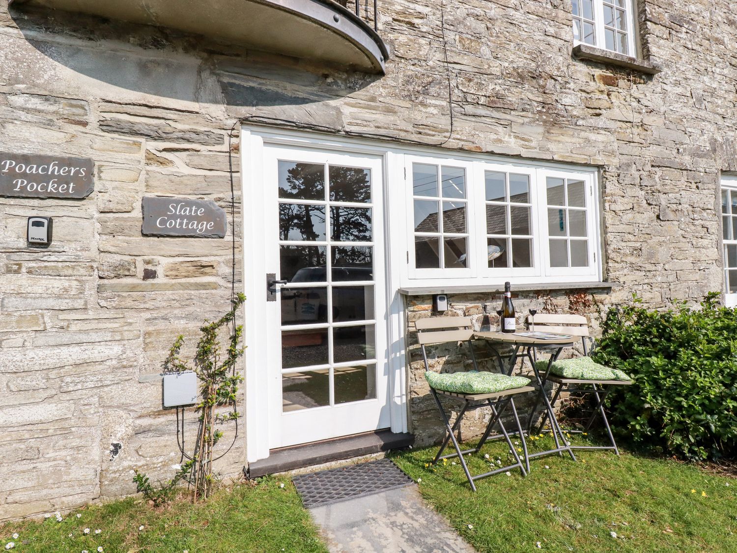 A stone cottage exterior with a white framed glass door and windows a small table with two chairs and cushions on grass at Slate Cottage Tredethy near Bodmin Moor