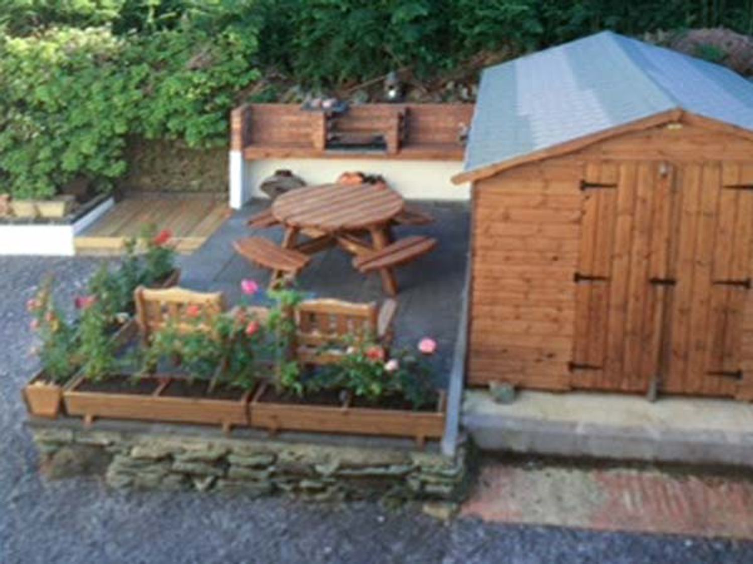 A garden patio area with a wooden picnic table and benches near a wooden shed with plants in flower beds at Glan Y Mor in Bull Bay