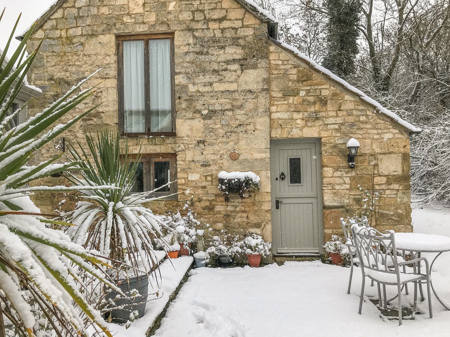 A stone house with a grey door and two windows in a snow-covered garden with metal table and chairs at The Old Cider House in Prestbury near Cheltenham