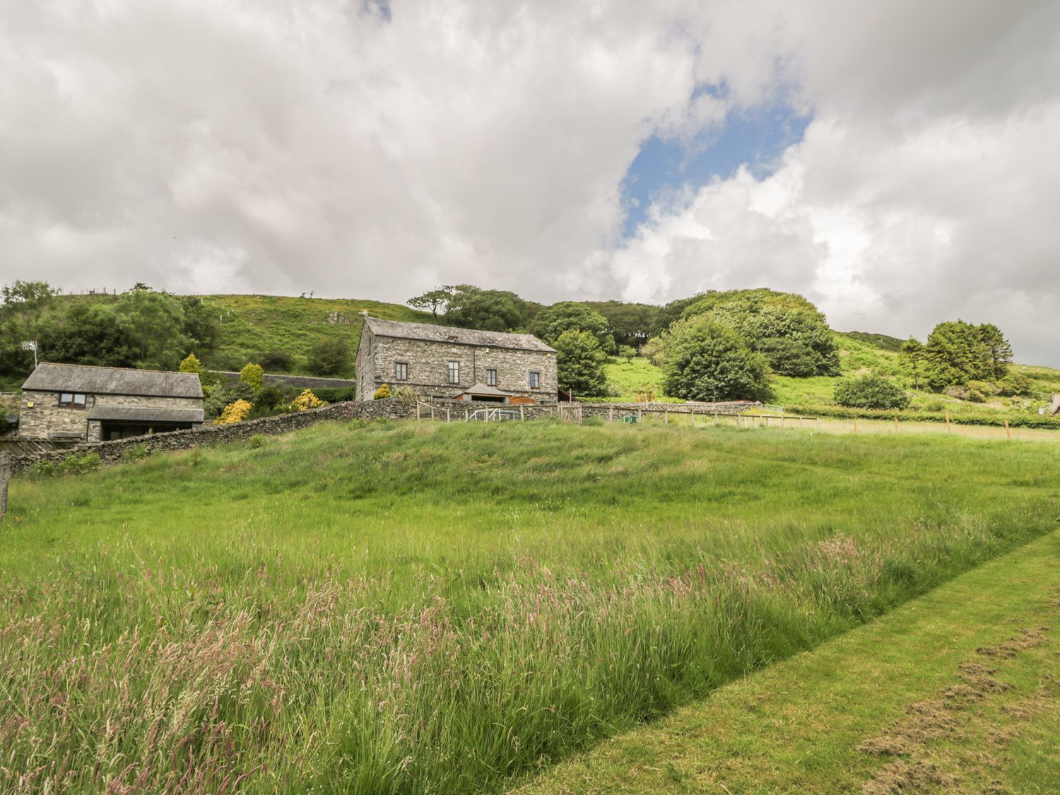 A grassy field with stone buildings and trees on a hill at Bank End Barn in Grizebeck