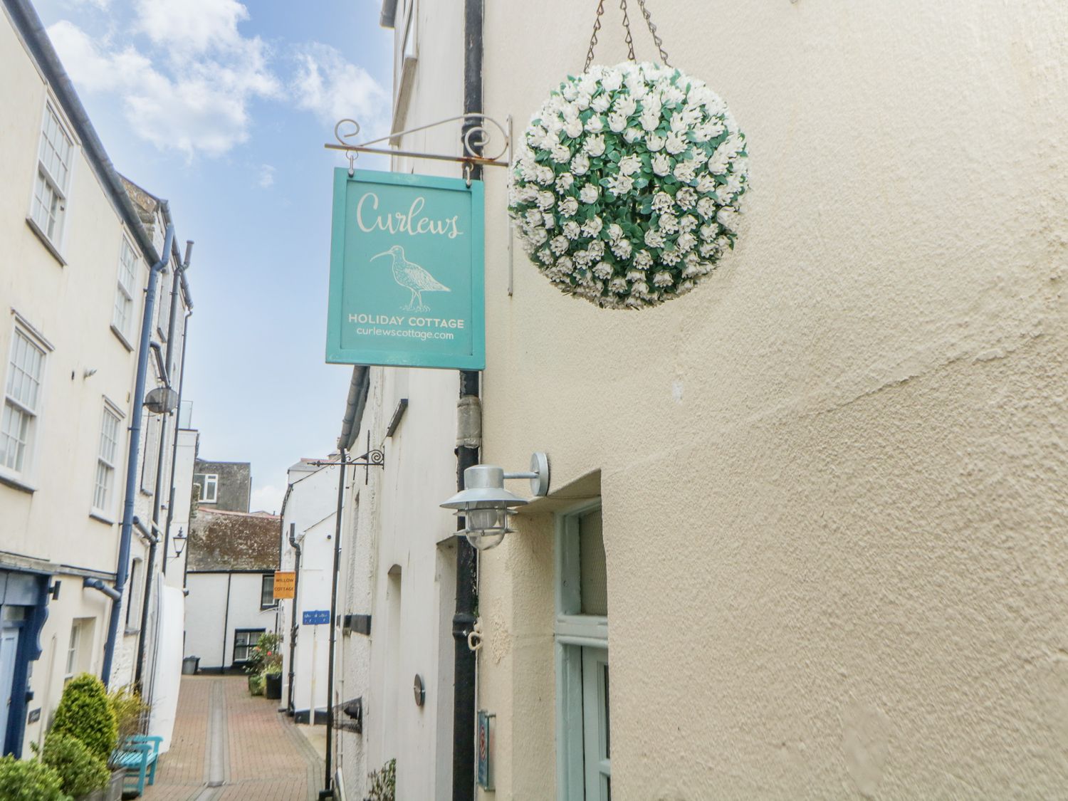 A narrow street with white buildings and a teal sign reading Curlews holiday cottage at Curlews Cottage in Looe