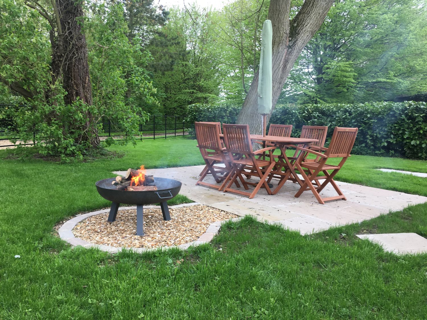An outdoor patio with wooden table and chairs near a fire pit with burning wood surrounded by grass and trees at The Dower House in Sharnbrook