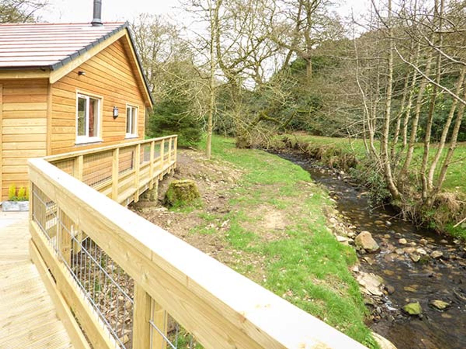 A wooden cabin with a railing deck next to a small stream and trees at Park Brook Retreat in Scorton