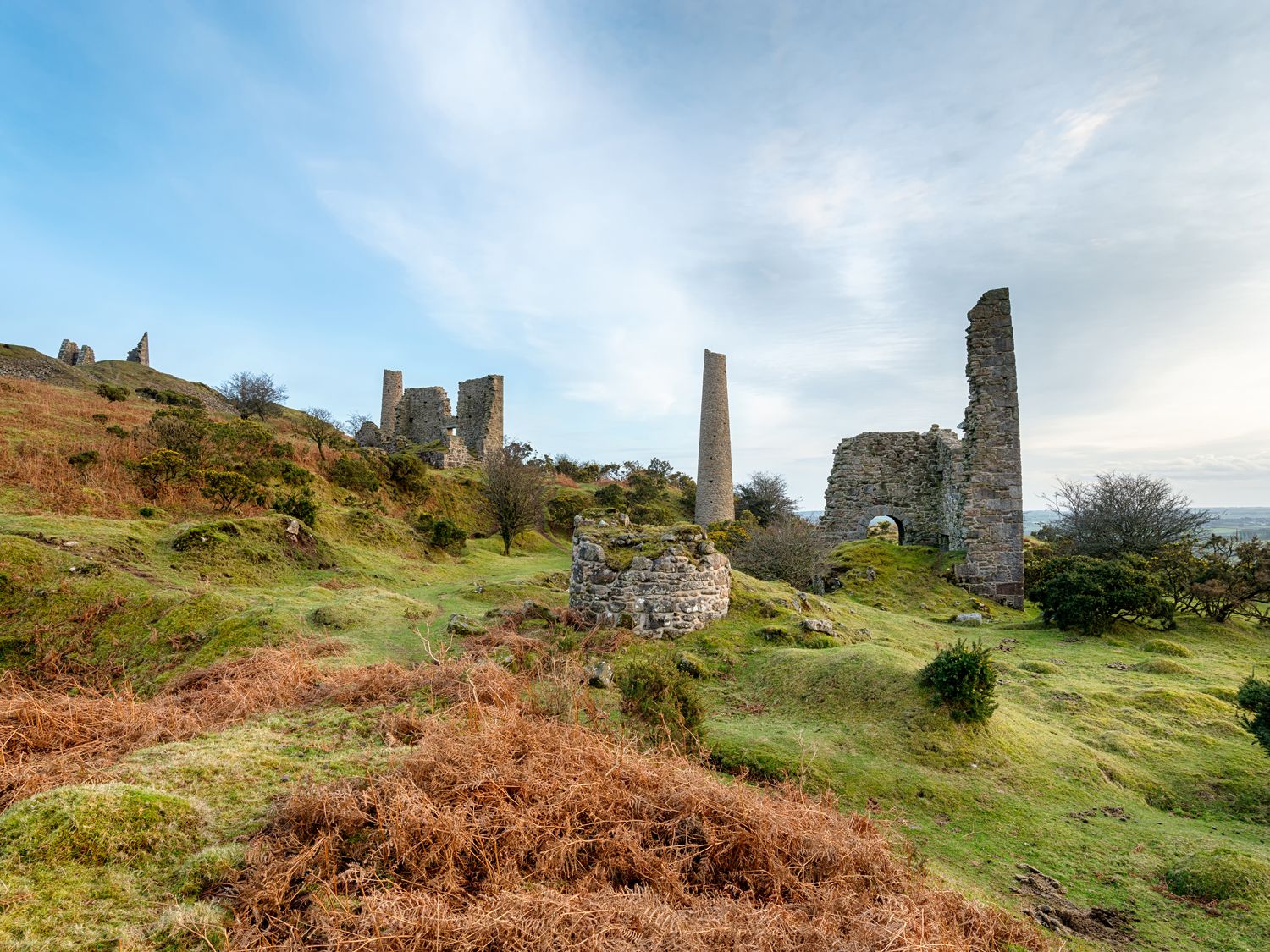 Ruins of stone buildings and towers on grassy hills with shrubs under a blue sky