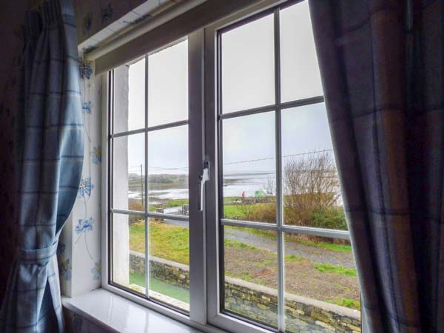 A window with checkered curtains showing a view of grass trees and water at No 1 Apt Brandy Harbour Cottage in Ballinderreen near Kinvara County Galway