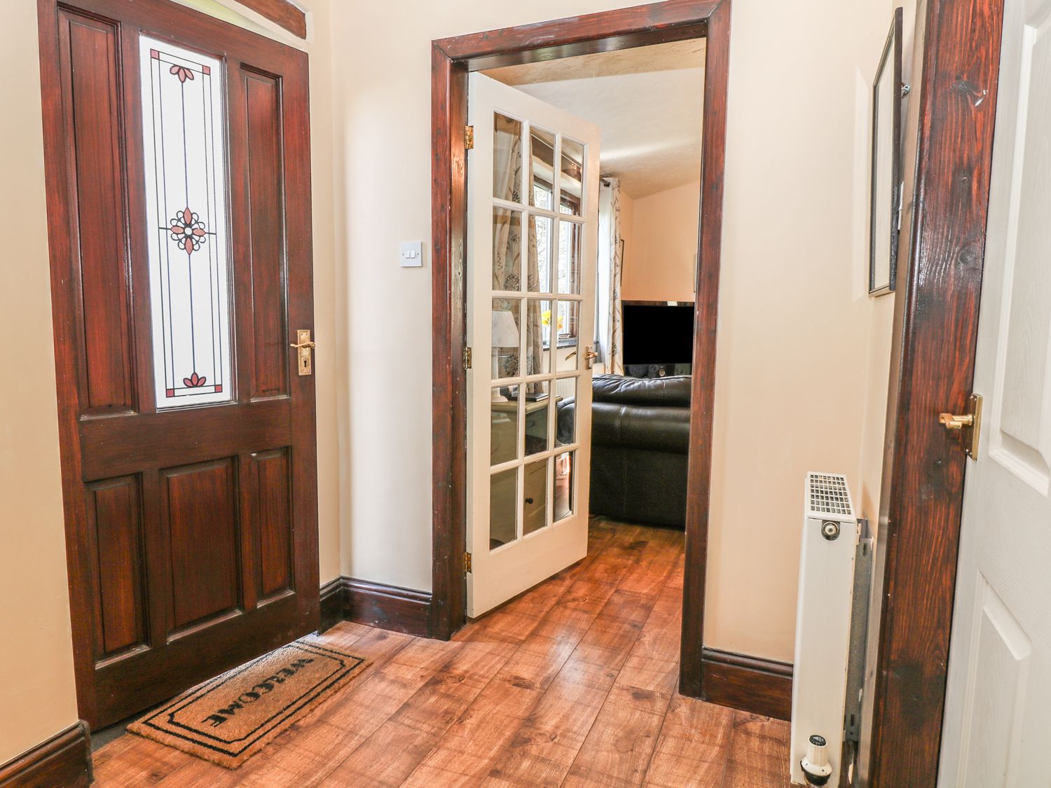 A hallway with a wooden front door and a doormat leading to a room with a glass paneled door and a visible black sofa at The Cottage at Moseley House Farm in Chinley