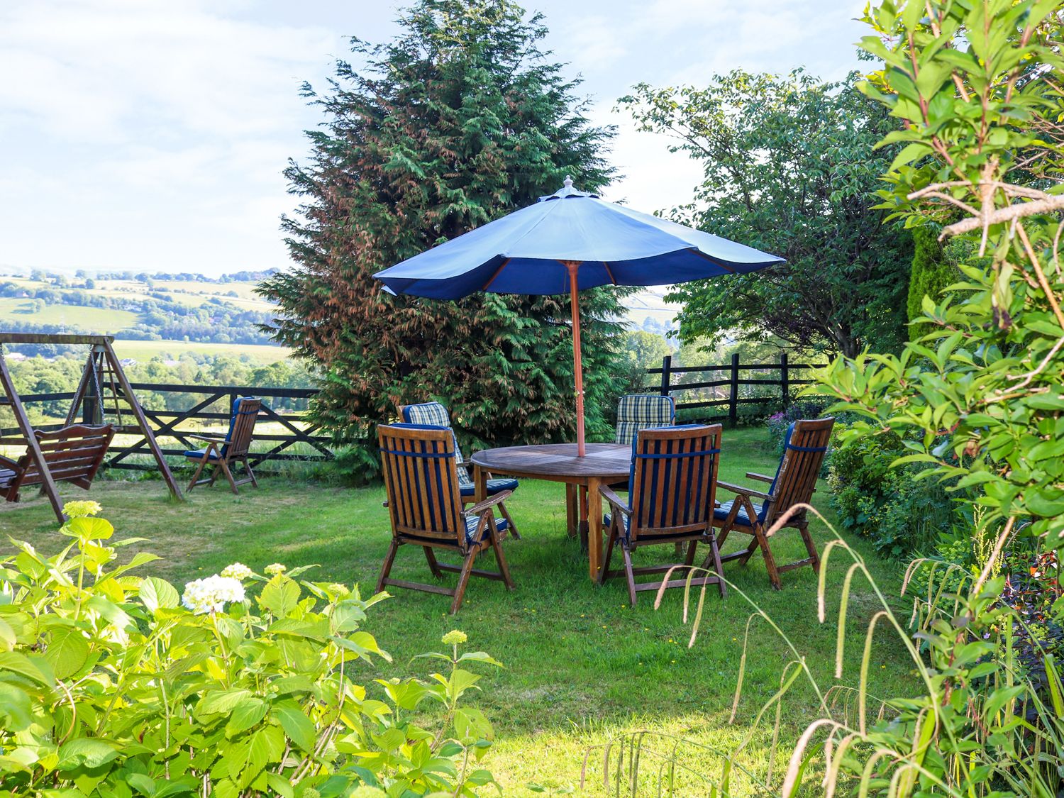 A garden with a round wooden table and chairs under a blue umbrella with plants and a wooden swing at The Cottage at Moseley House Farm in Chinley