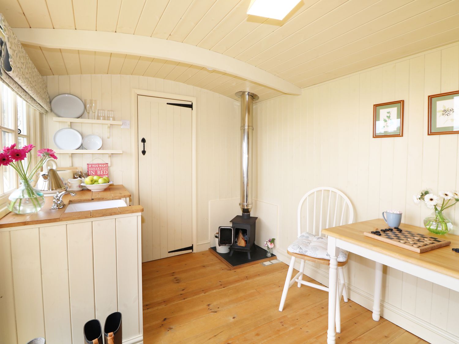 A small kitchen area with a wood stove a table and chair with a game board and flowers at Willow Burlawn near Wadebridge