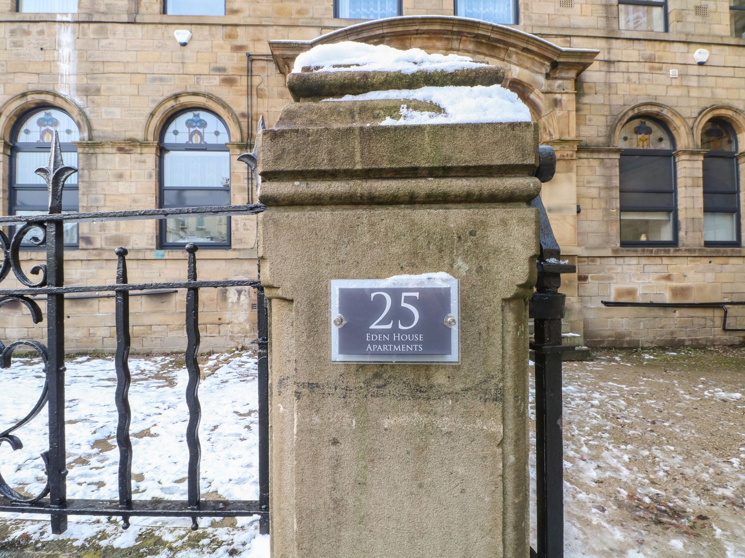 Stone pillar with a sign reading 25 Eden House Apartments near a black metal fence and a building with arched windows
