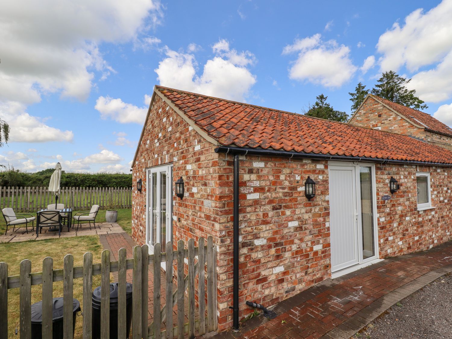 A brick single-story building with a tiled roof and white doors and windows in a fenced yard with outdoor seating at Westerby Lodge in Alford