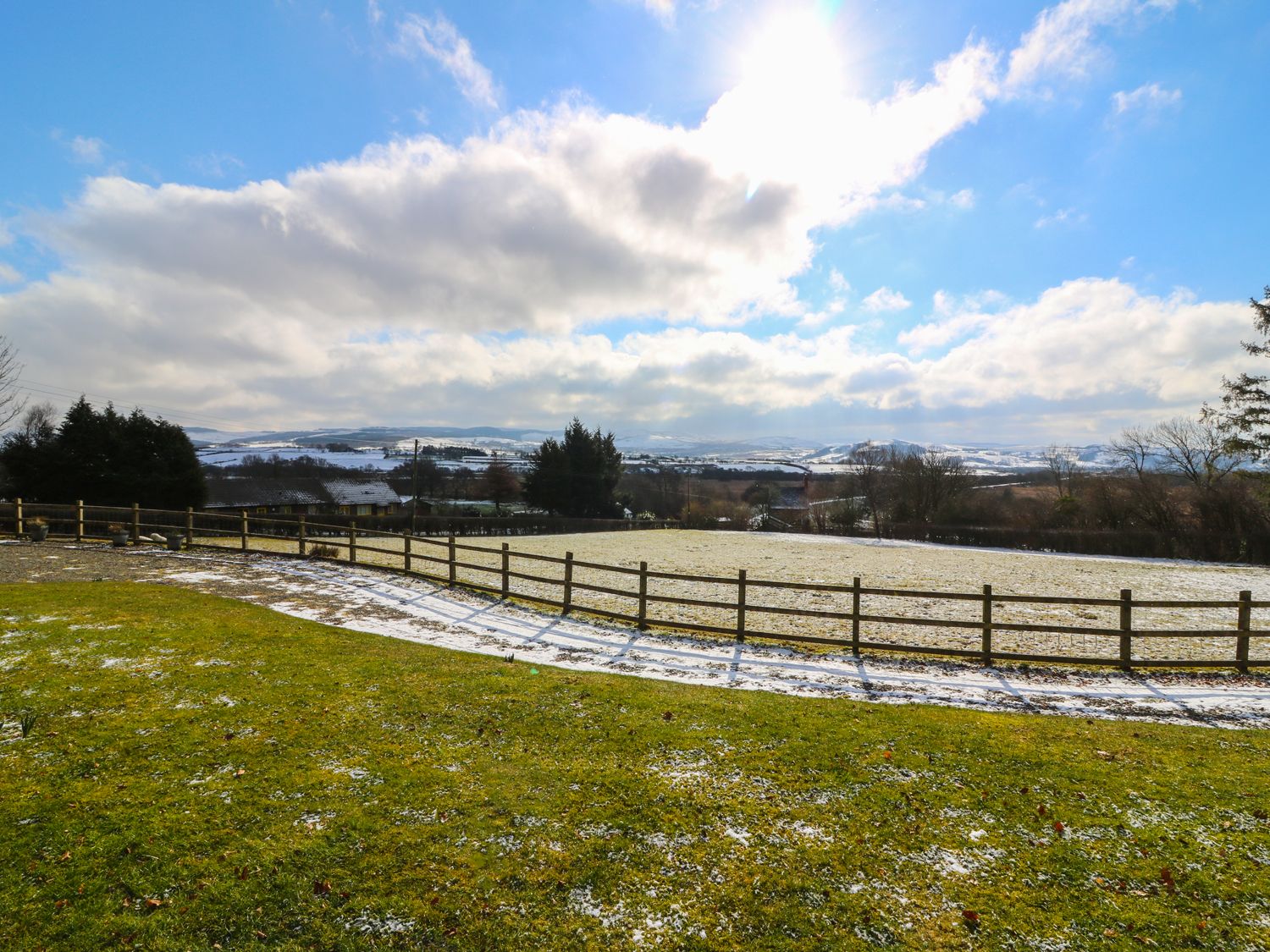 A fenced grassy field with patches of snow and distant houses under a partly cloudy sky at Dolau Meurig in Pontrhydfendigaid