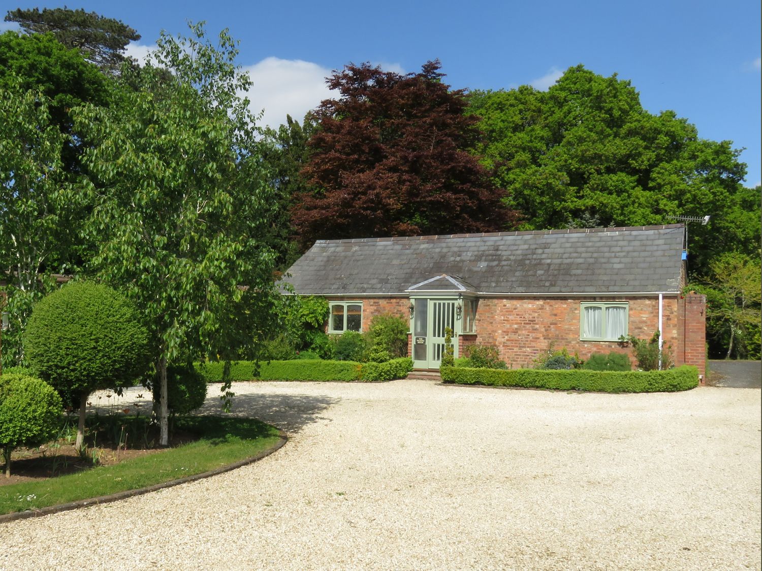 A small brick house with a gravel driveway and various green trees and bushes at The Packing House Hanley Castle near Malvern