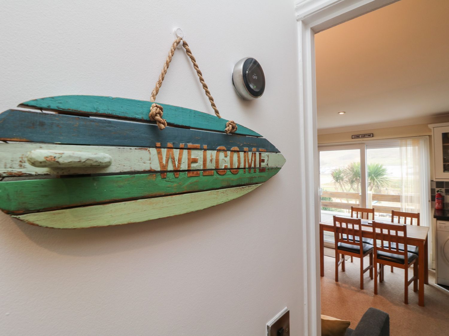 A wooden welcome sign hanging on a wall next to a thermostat with a dining area and sliding doors overlooking outdoor plants at Ocean Blue in Porthtowan