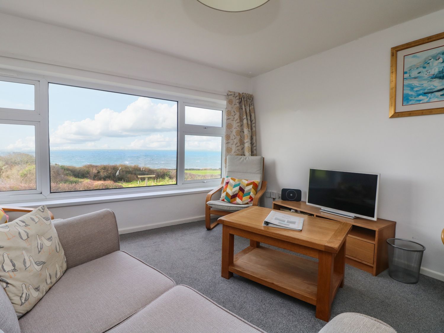 A living room with a large window overlooking the sea a sofa armchair wooden coffee table and television at The Barn in Gunwalloe