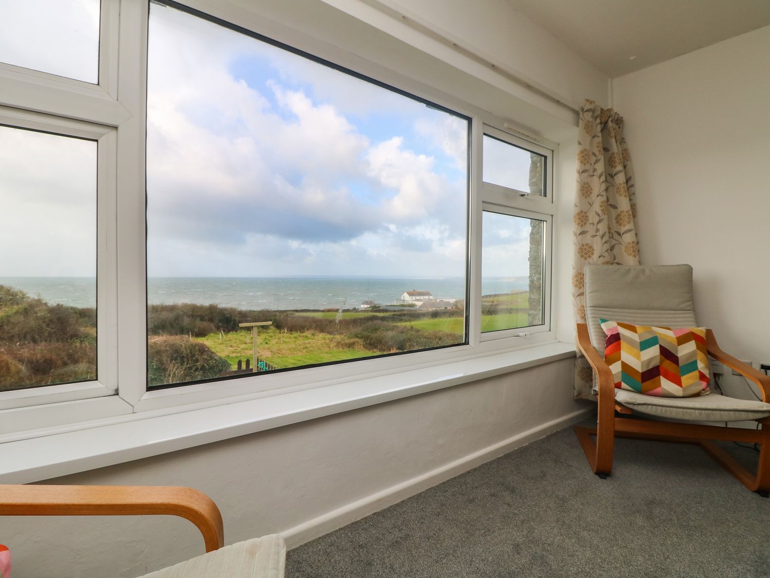A room with a large window overlooking a grassy field and the sea with a chair and colorful cushion at The Barn in Gunwalloe