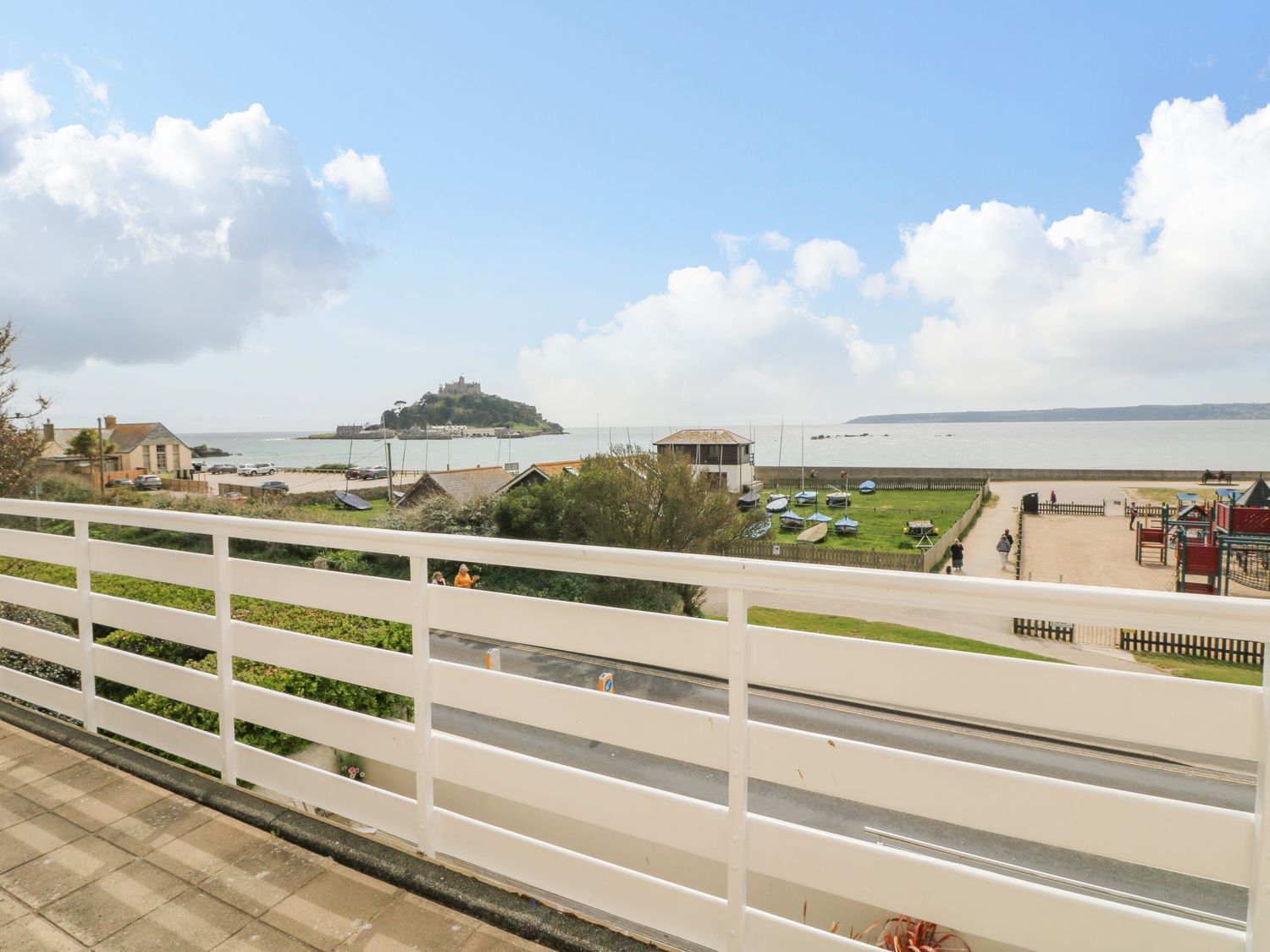 A balcony overlooking a road green area with boats playground and a sea with an island at Sea Breeze in Marazion