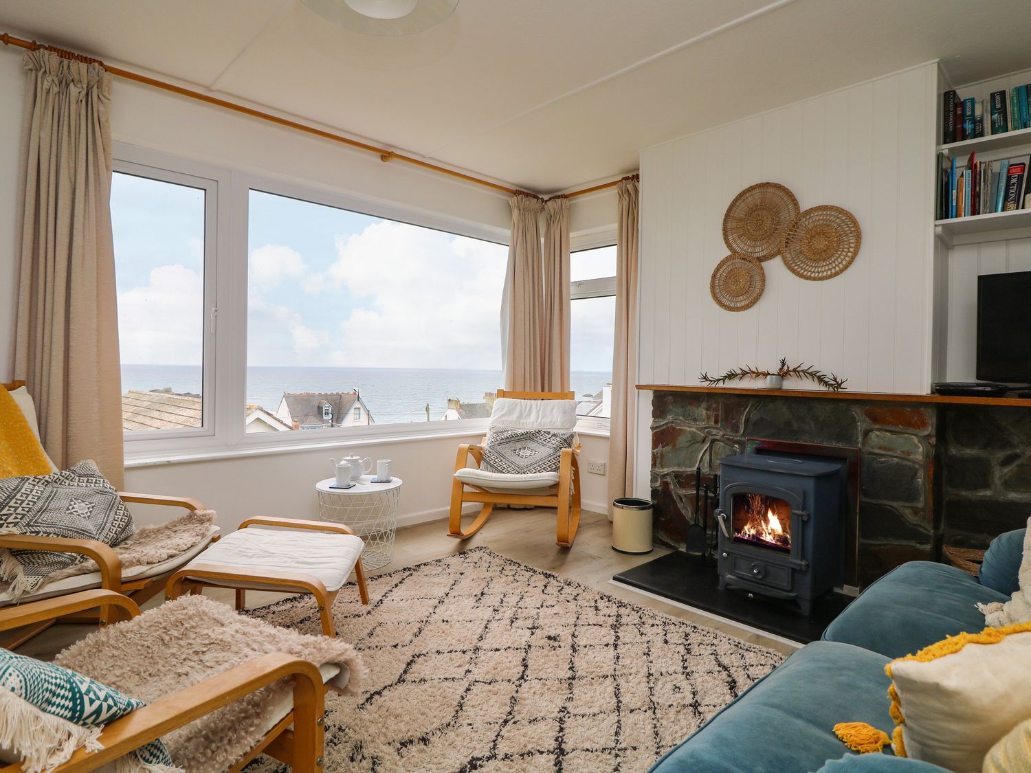A living room with wooden chairs, a blue sofa, a rug, a stove fire, and a window with a sea view at Wootton Gray in Mousehole