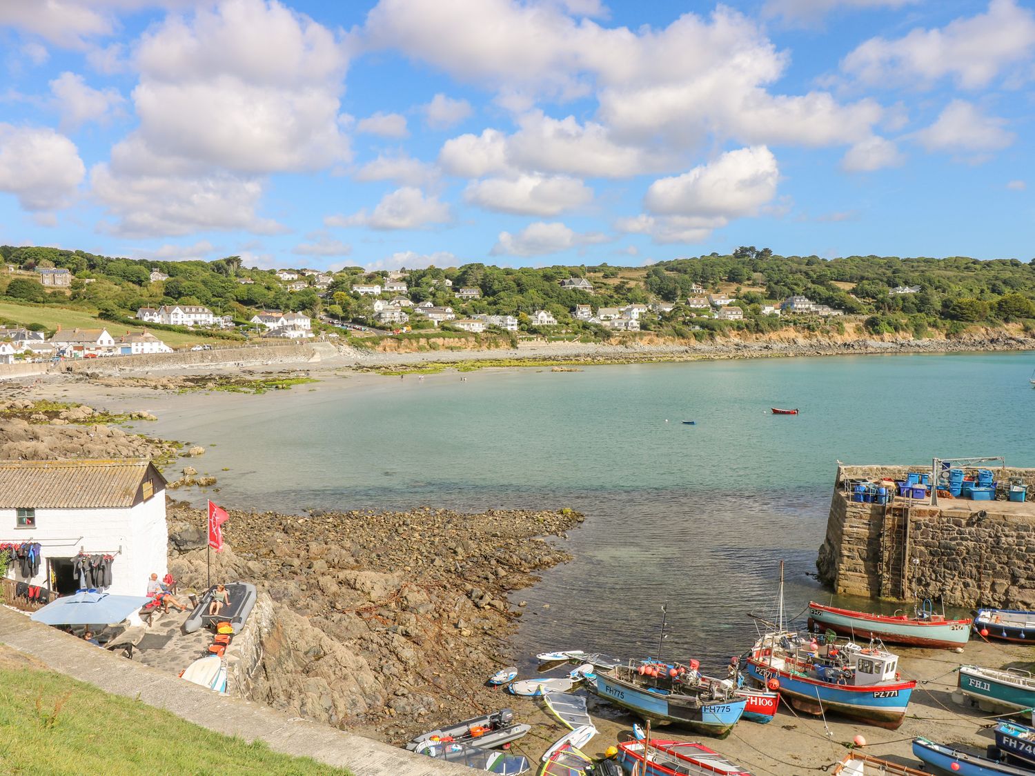 A coastal harbor with small boats docked near a stone pier and houses on a hillside at Eden House Wing in St Keverne
