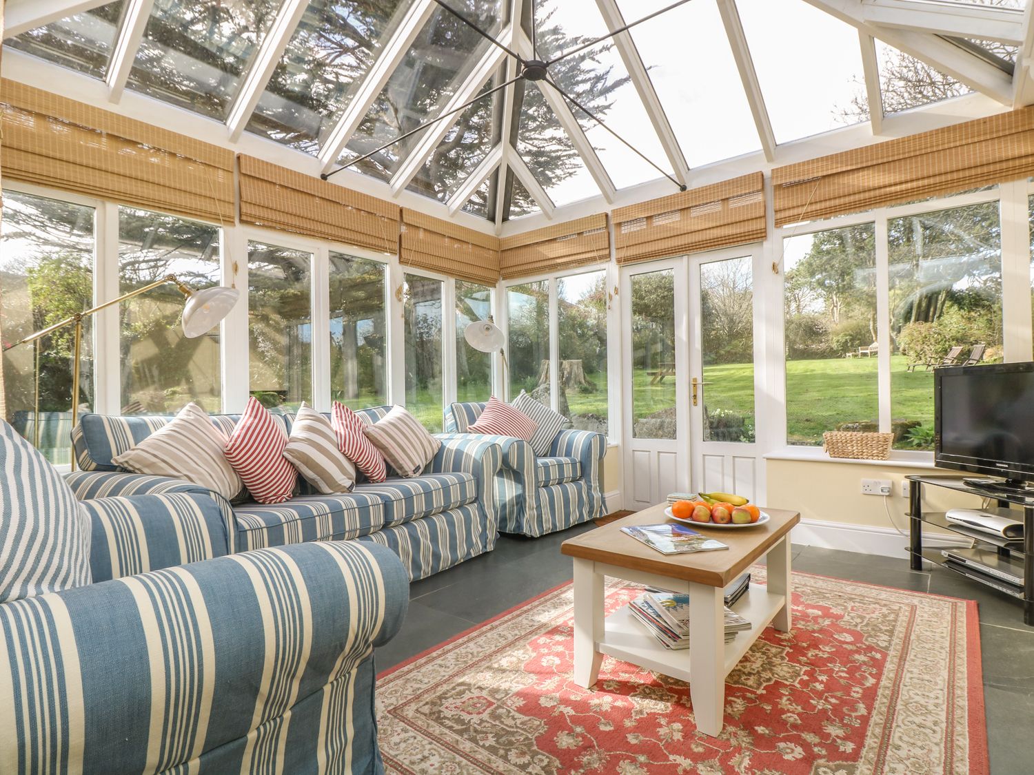 A sunroom with striped sofas, a coffee table with fruit on a rug, and a television at Boskednan Wartha in Penzance