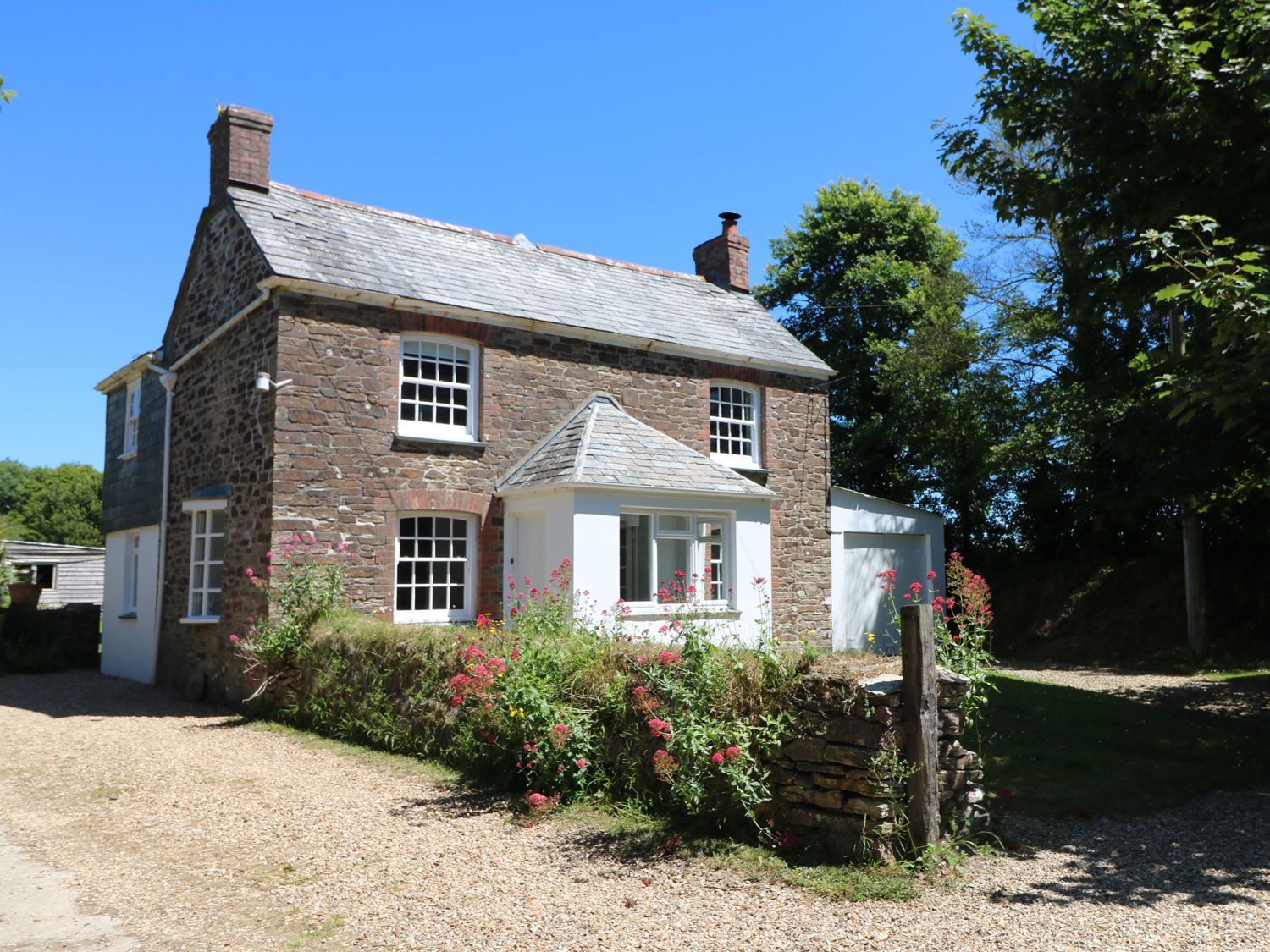A stone house with white windows and a small porch surrounded by flowers and trees at Trenouth Cottage in Padstow