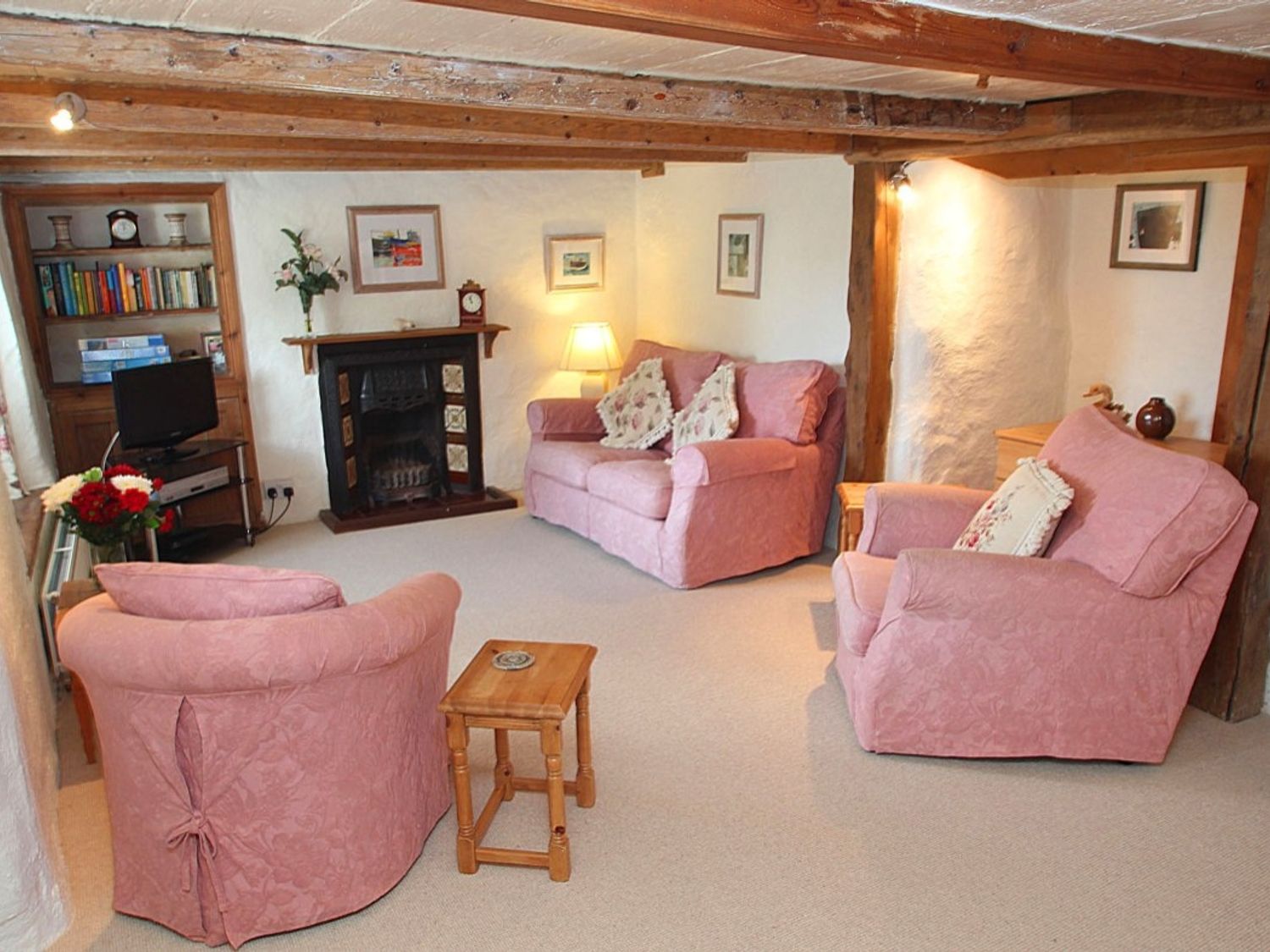 A living room with exposed wooden beams pink upholstered sofa and chairs a fireplace and wooden furniture at Rose Cottage in Manaccan