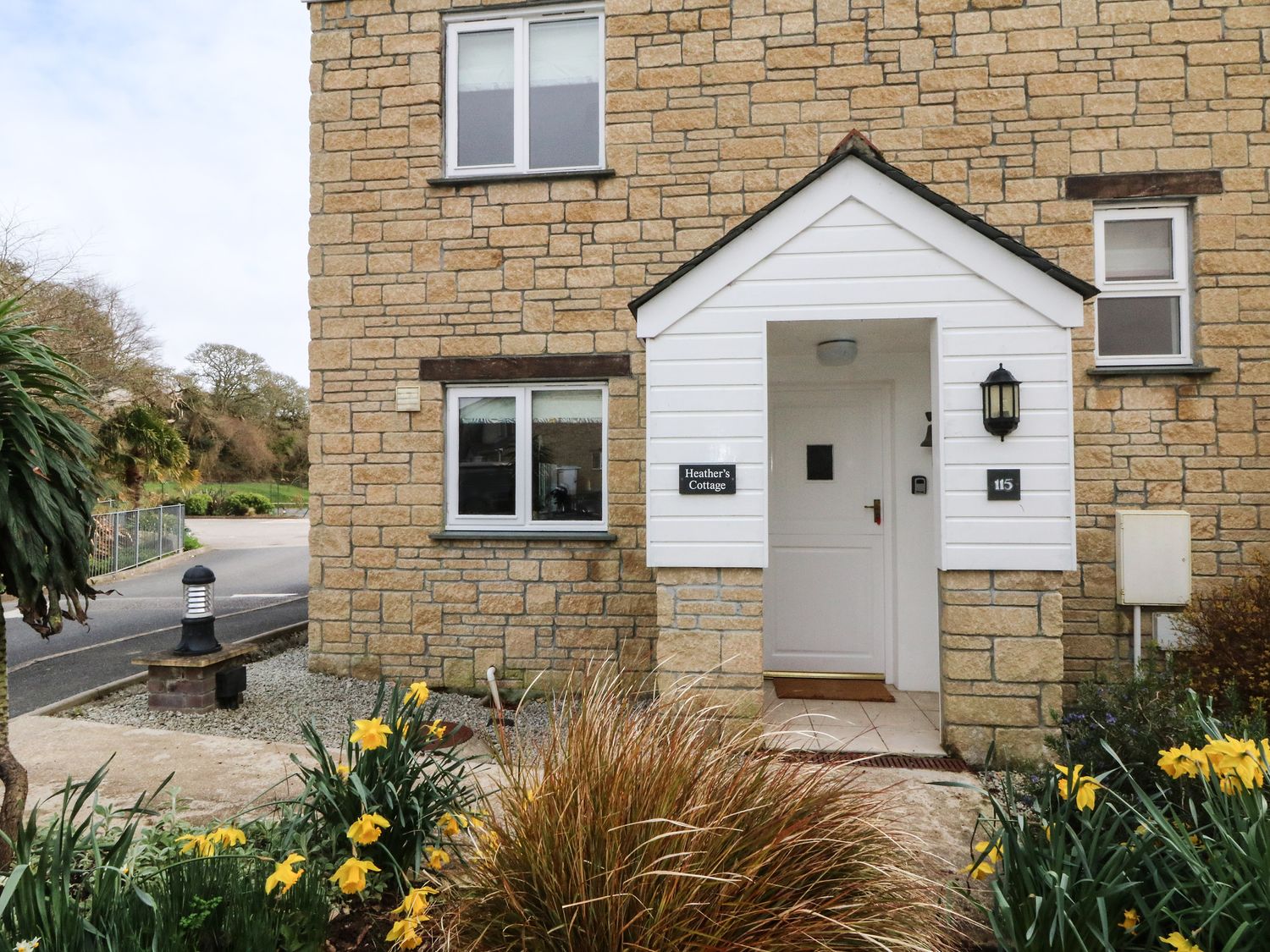The entrance of a stone building with a white door and porch surrounded by plants and flowers at Heathers Cottage in Falmouth