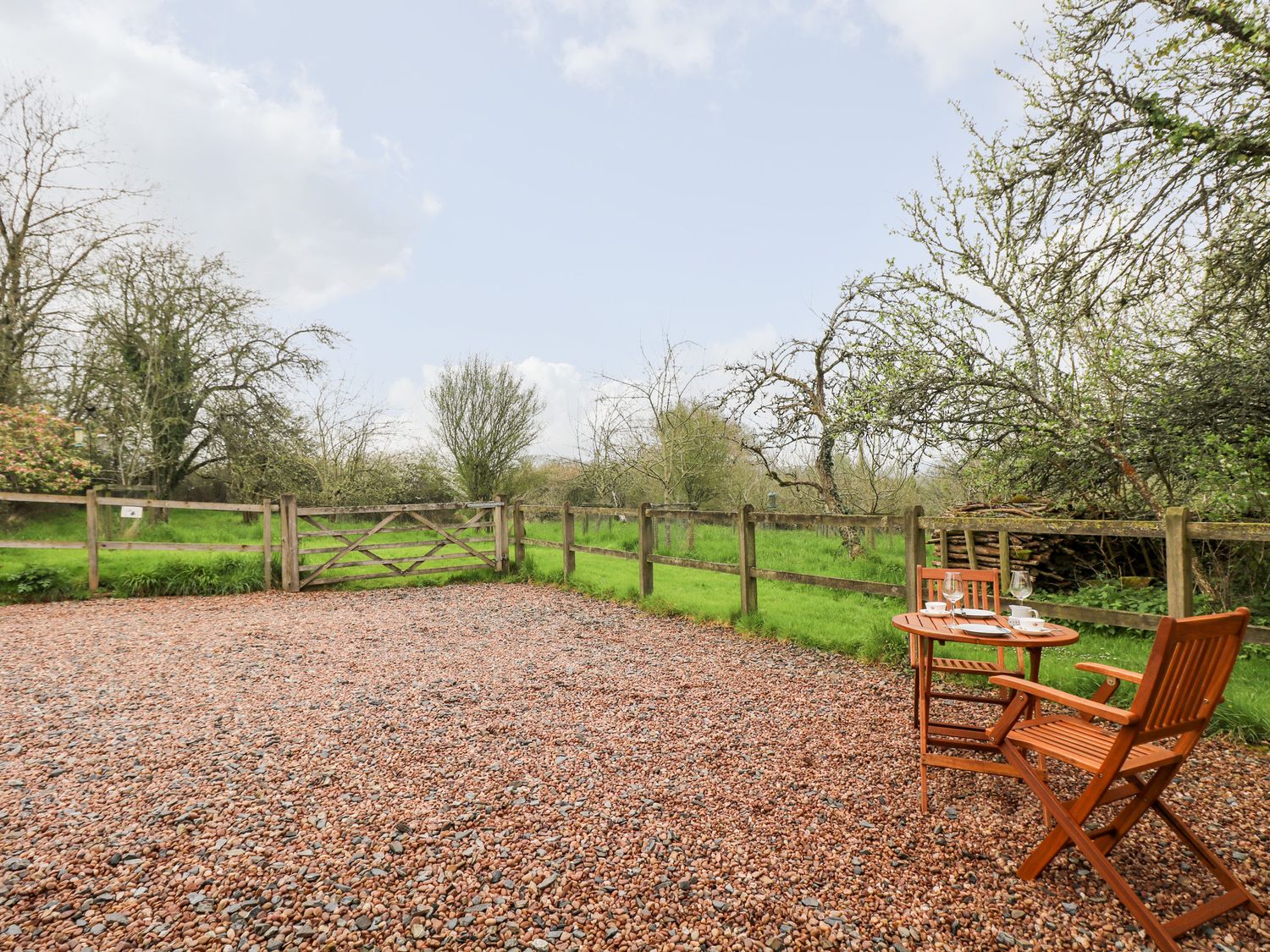 A gravel area with a wooden table and two chairs set with glasses and cups beside a wooden fence in a green garden at Plum Cottage in Colebrooke near Crediton