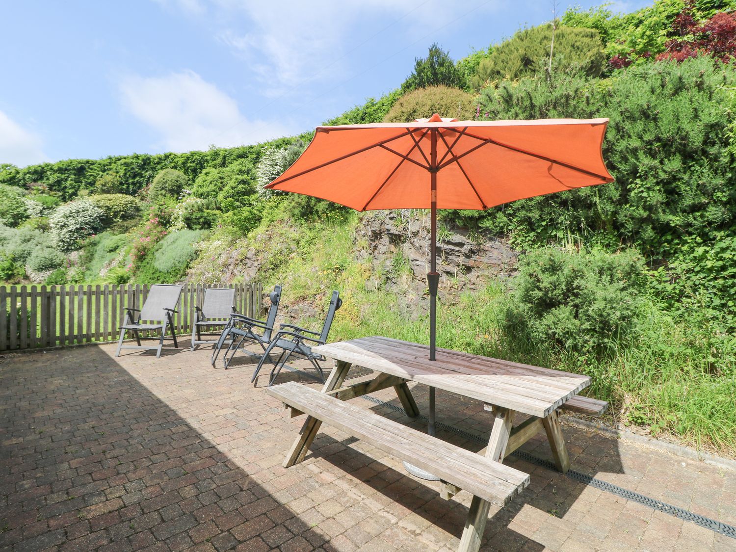 An outdoor patio with a wooden picnic table and orange umbrella surrounded by garden chairs and greenery at Roosters in Lostwithiel