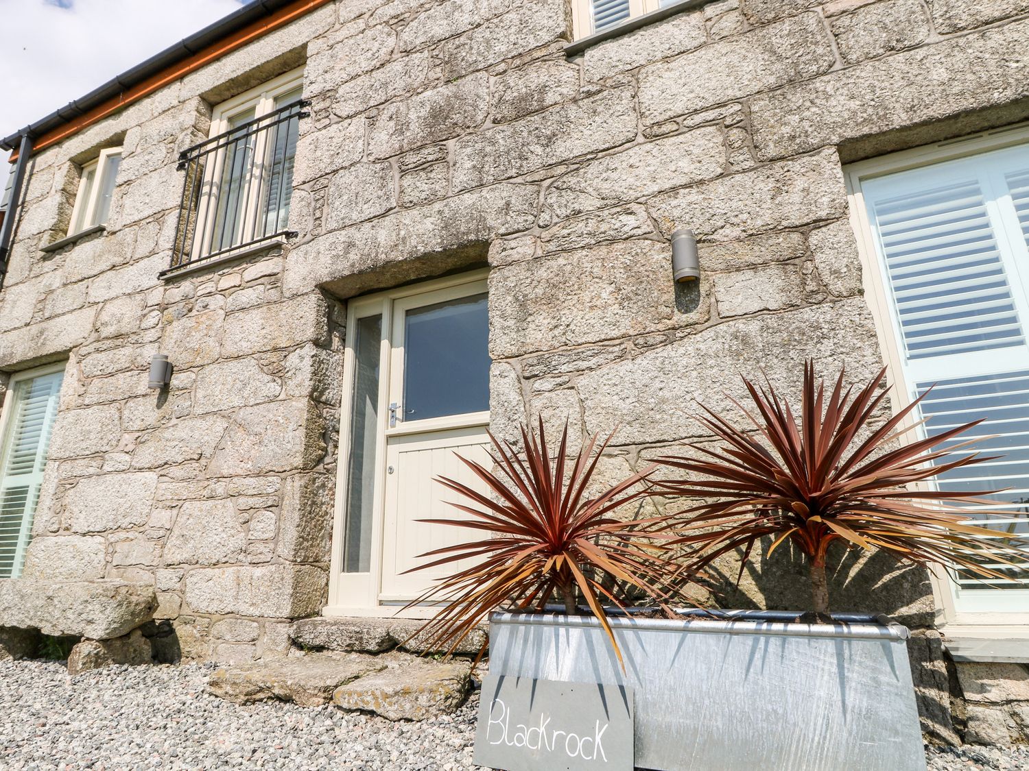 The exterior of a stone building with a cream-colored door and windows with shutters and a metal planter with two spiky plants and a sign that reads Blackrock at Black Rock in St Austell