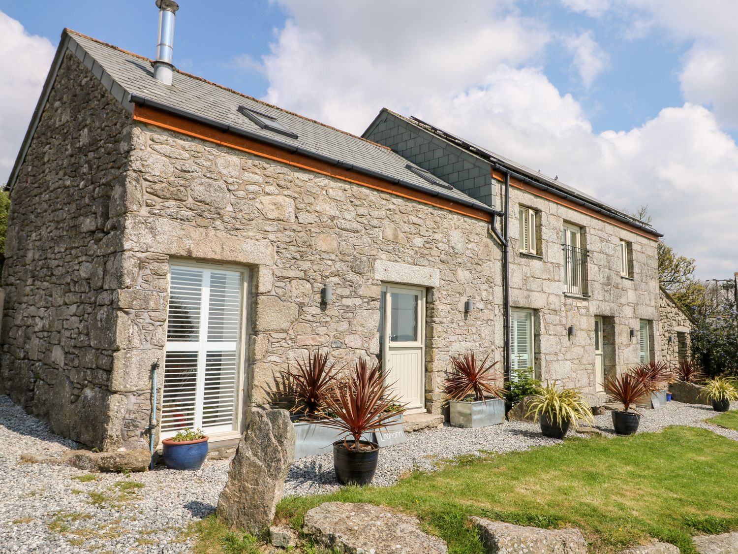 A stone house with multiple windows and potted plants outside on a gravel and grass area at Leghorn in St Austell