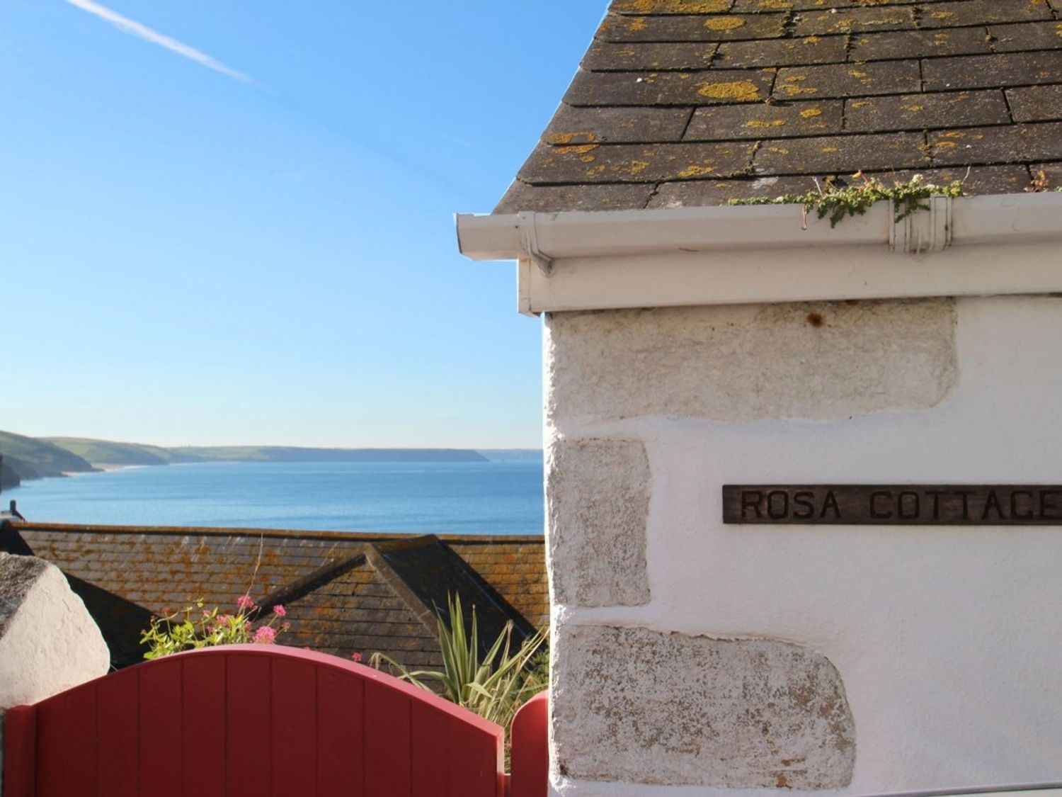 A view of the sea with rooftops and a red gate in front of a white stone building with a sign that says Rosa Cottage in Porthleven