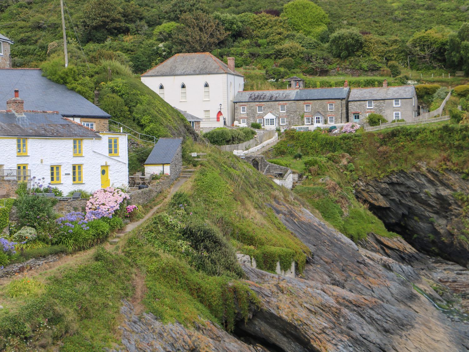 Cottages on a hillside with flower gardens and rocky coastline at Kerbenetty Harbour Cottage in Portloe