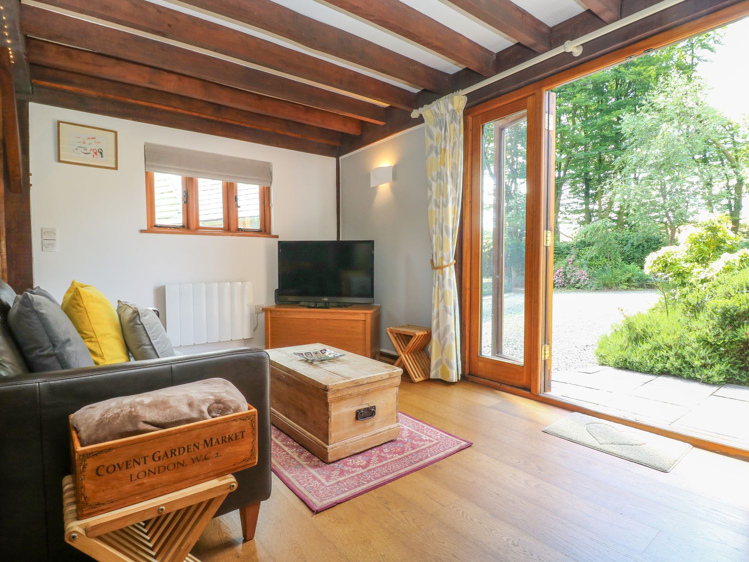 A living room with wooden beams on the ceiling a leather sofa cushions a wooden chest as coffee table a television on a wooden cabinet and glass doors opening to a garden at Oaklands Near Boscastle
