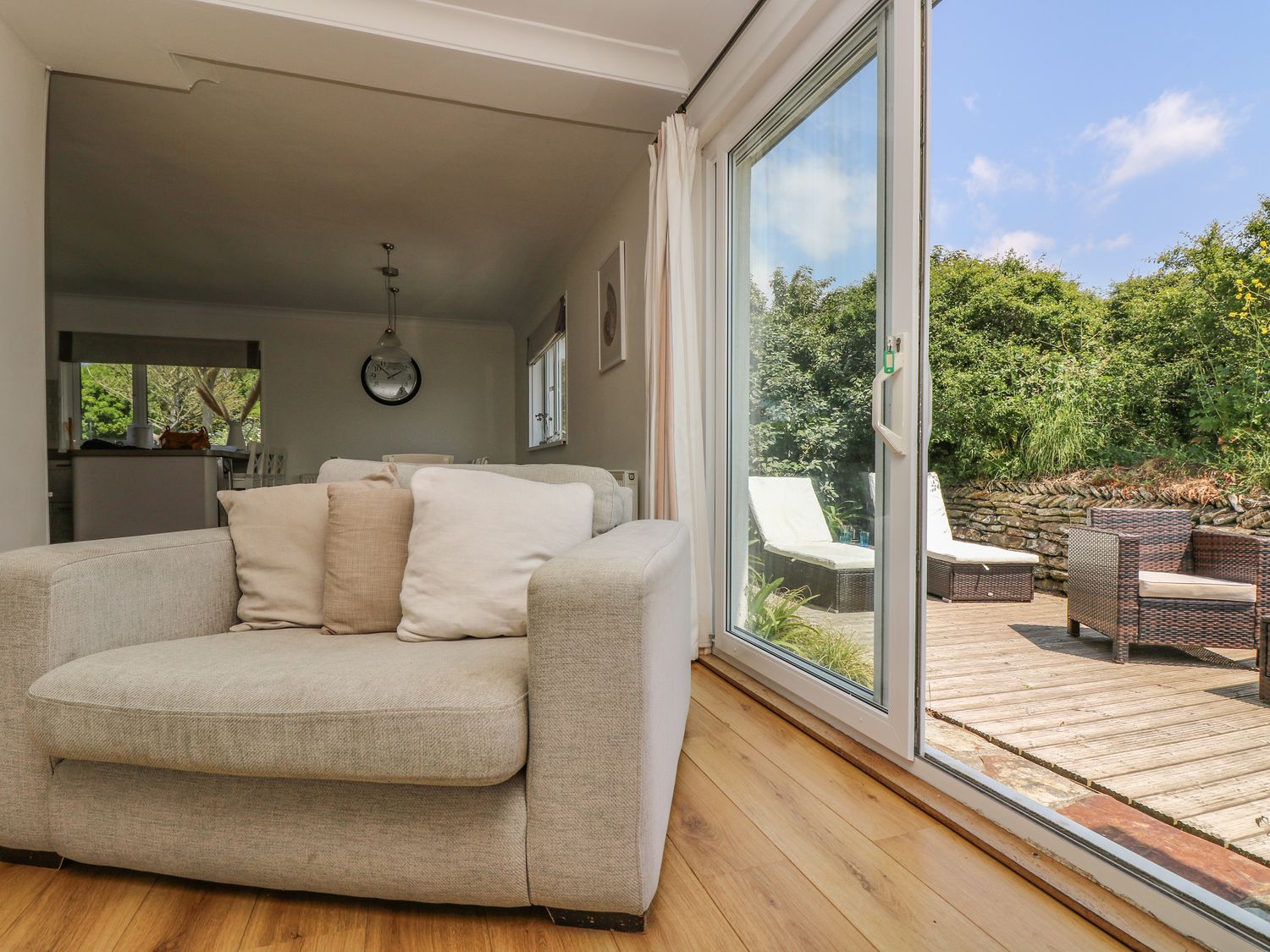 A living room with a sofa and cushions next to sliding glass doors leading to a wooden outdoor deck with patio furniture at Seaport Lodge in Mawgan Porth