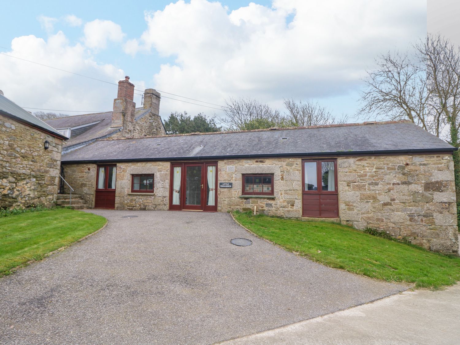 A stone building with three red doors and two windows with grass areas on either side at Little Tregarrick in Helston