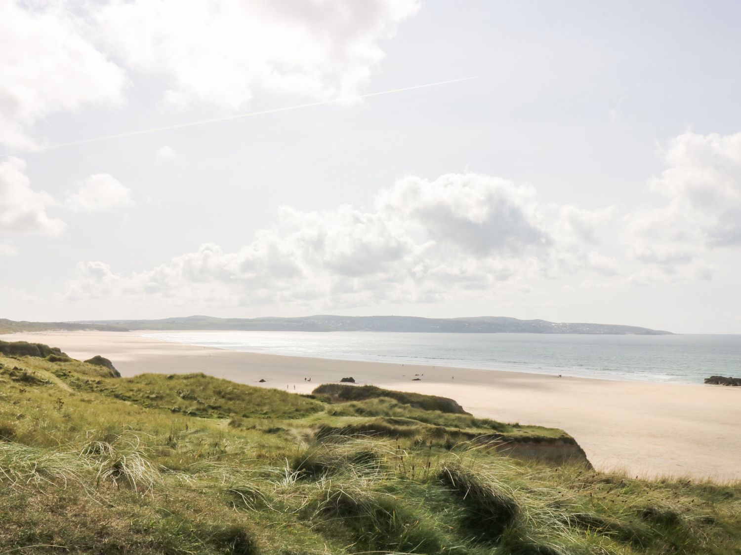 A sandy beach with people near the shoreline and grassy dunes in the foreground at Pentewan in Hayle
