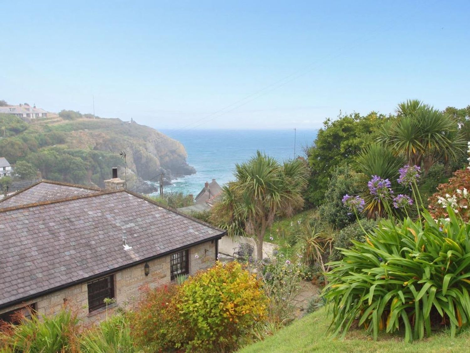 A garden with various plants and a bench overlooking a stone house roof and the ocean at Mengarth in Cadgwith