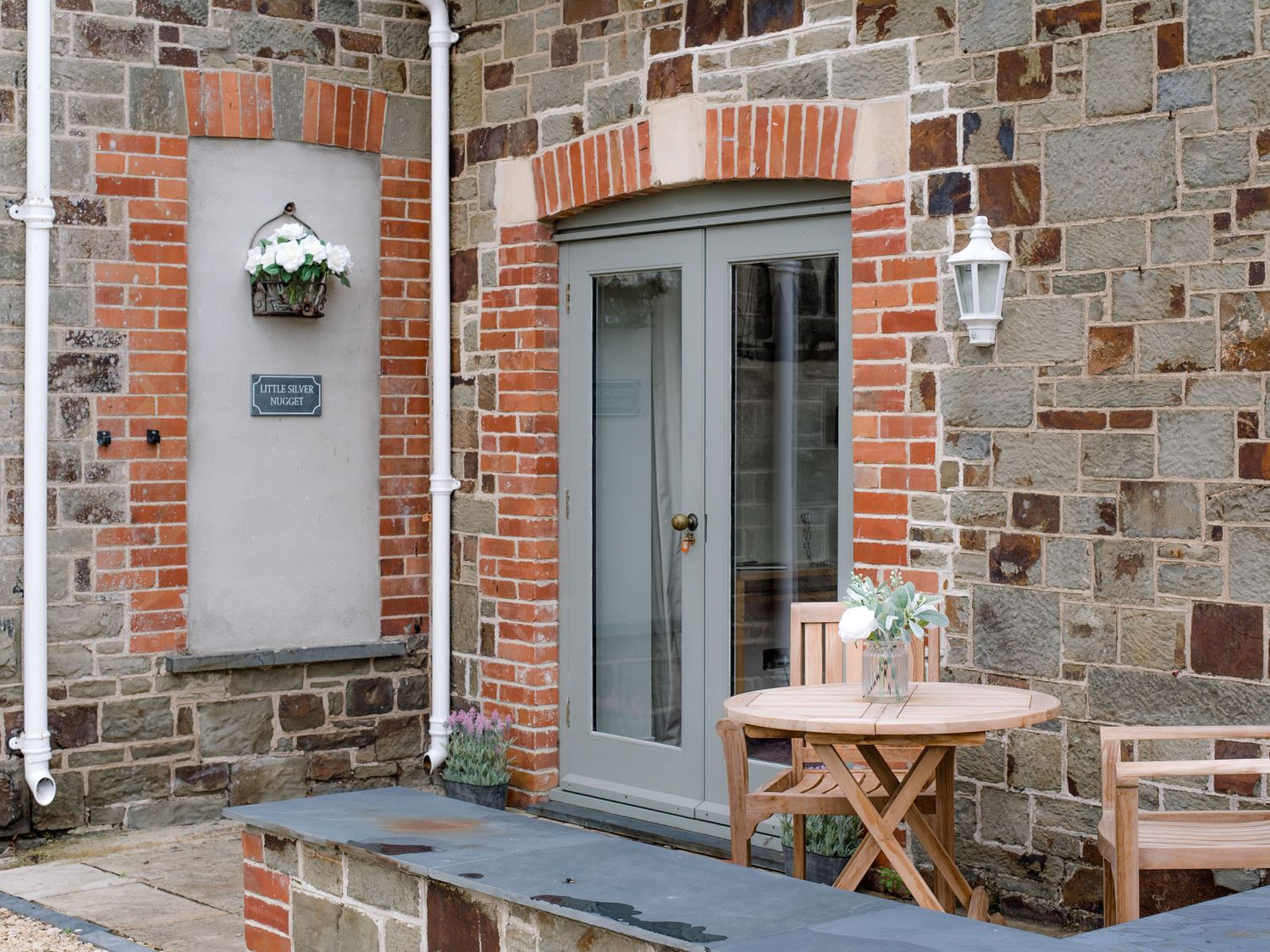 A stone wall with a gray door and a round wooden table with chairs and flowers at Little Silver Nugget in Umberleigh