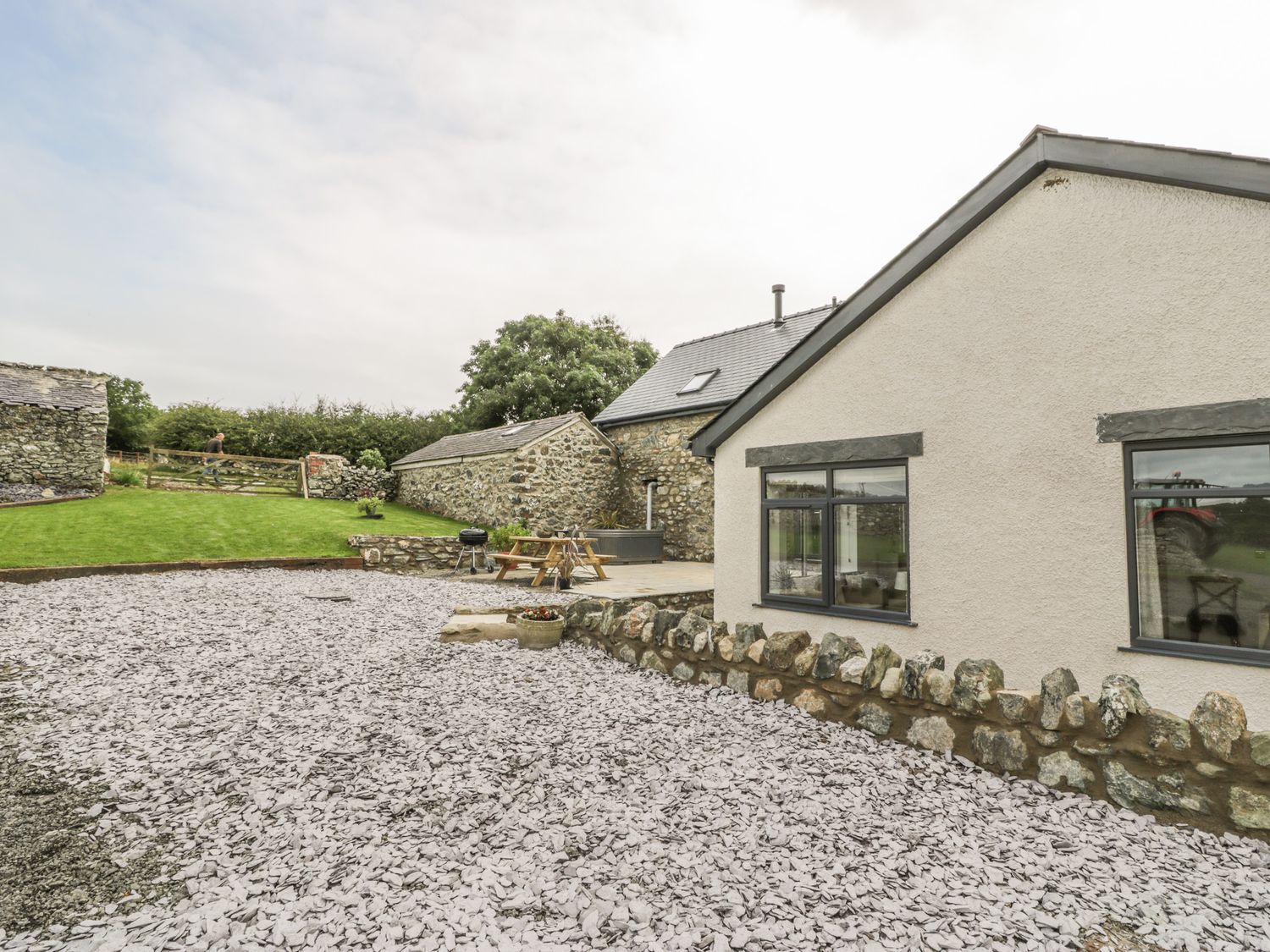 An outdoor area with a gravel driveway a stone wall a patio with a picnic table and a charcoal grill adjacent to a house and stone buildings at Storws in Pentraeth