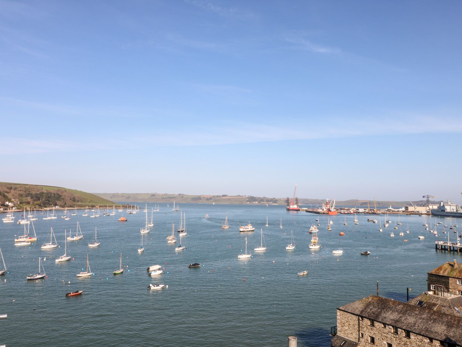 A harbor with many sailboats and ships on the water near industrial docks and a coastline at Oakwood Treesmill near Lostwithiel