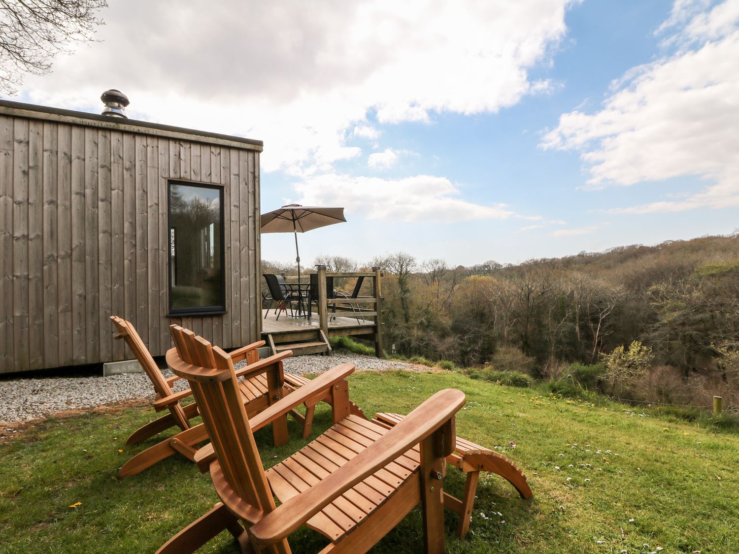 Wooden chairs on grass near a wooden cabin with a deck and umbrella overlooking a forested area at Oakwood Treesmill near Lostwithiel